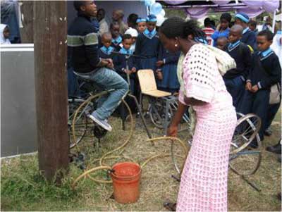 Photo of a man riding a bicycle mounted on a stand, with attached pump and hose assembly.