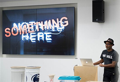 A man stands behind a lecturn with a video screen that reads "Something Here."